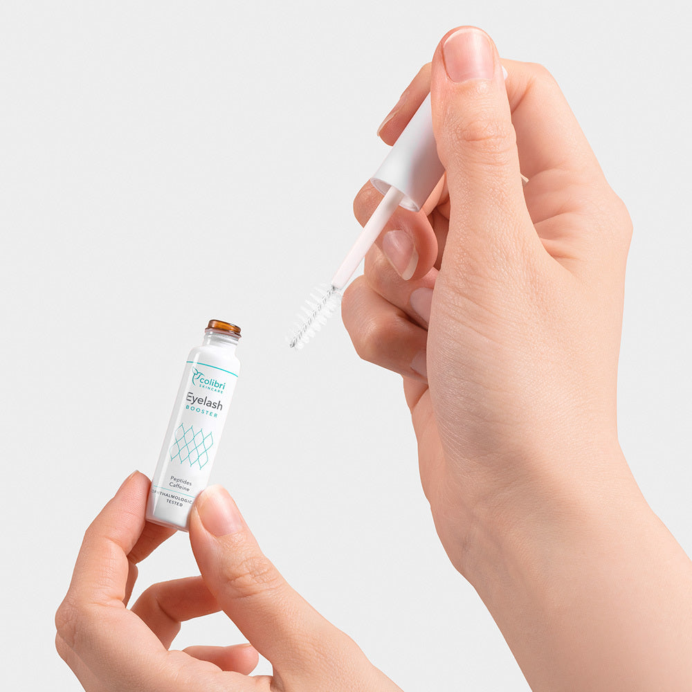 Hände halten einen geöffneten Flakon einer Wimperntusche mit einer Bürste gegen einen weißen Hintergrund.