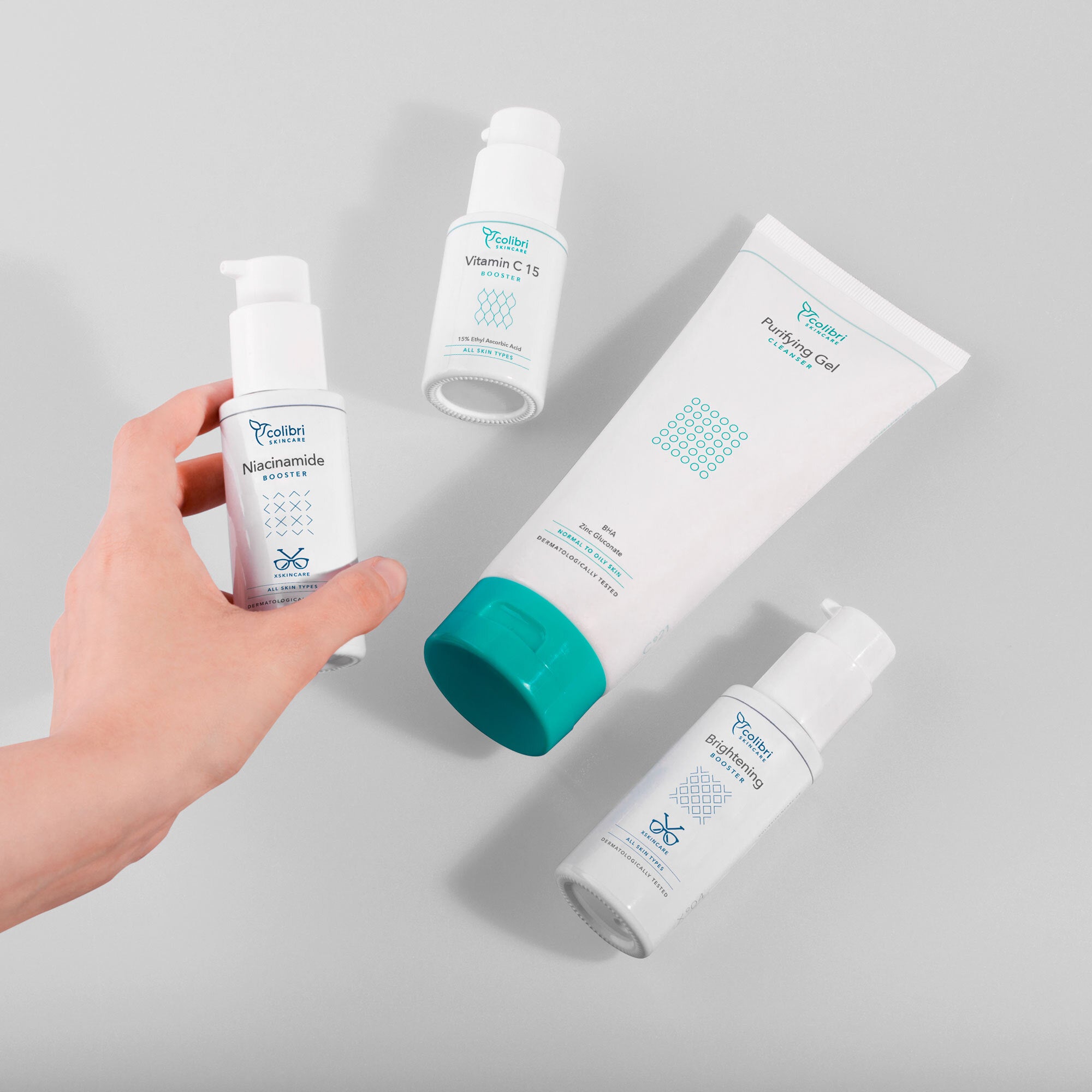 Eine Hand hält ein Fläschchen mit Niacinamide-Boostern. Daneben liegen drei weitere Hautpflegeprodukte auf einem grauen Hintergrund.