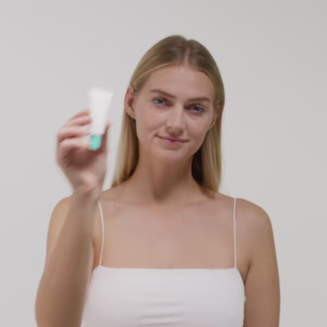 Junge Frau in weißem Top hält eine kleine Tube vor neutralem Hintergrund.