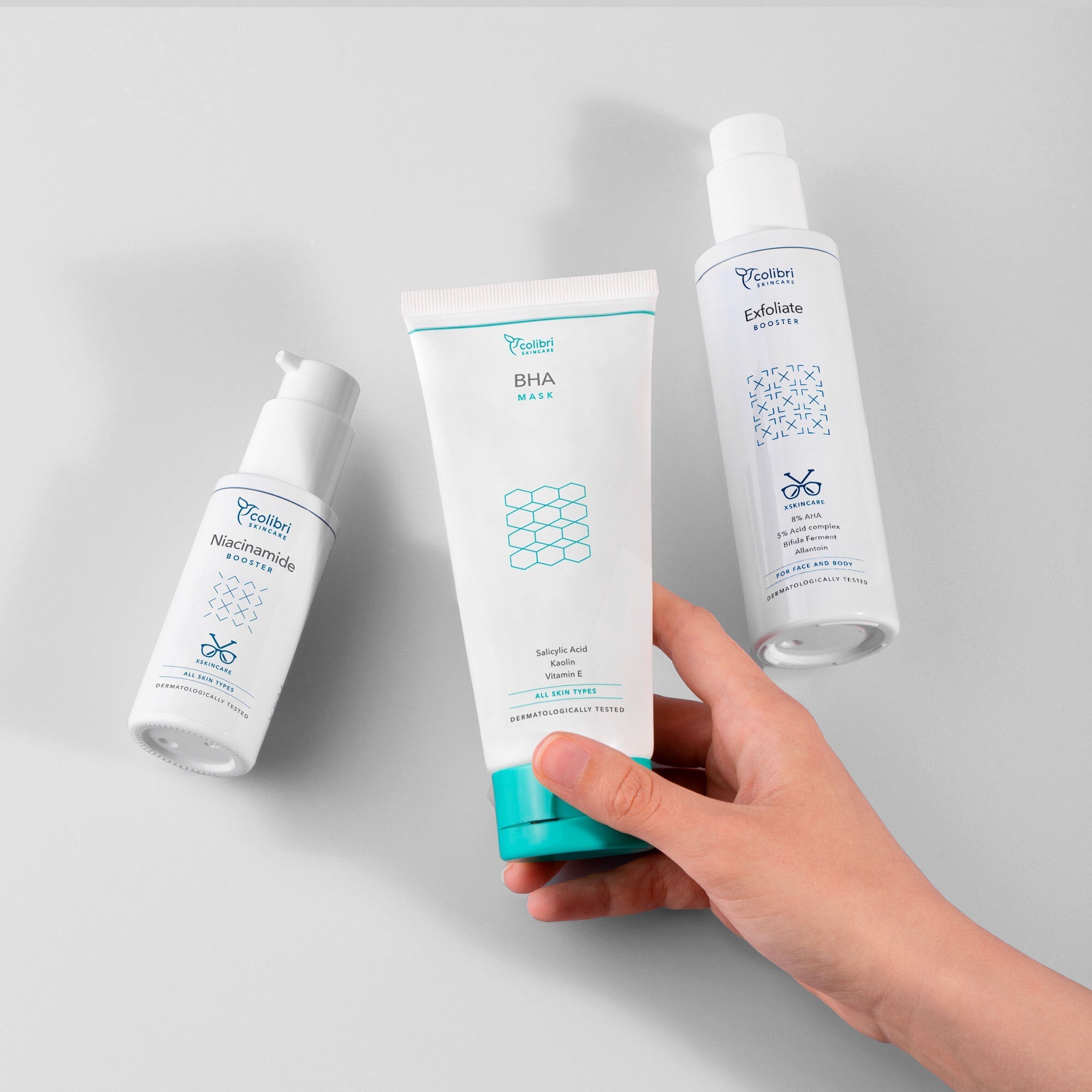 Drei Hautpflegeprodukte auf grauem Hintergrund, eine Hand hält eine BHA-Maske, daneben Niacinamide und Exfoliate Booster.