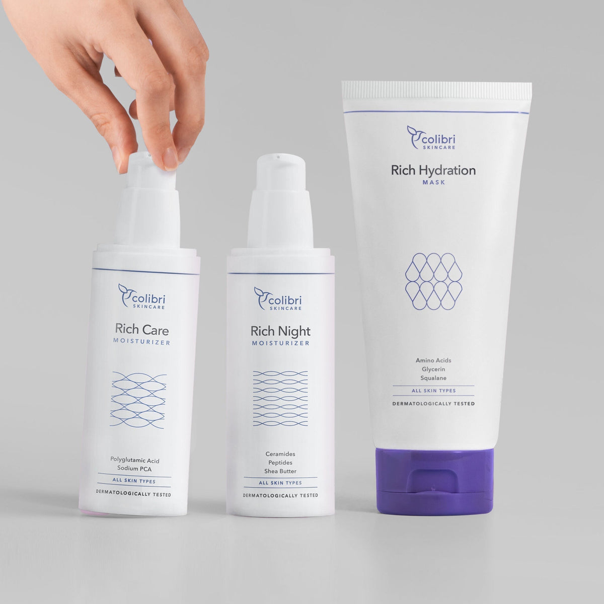 Eine Hand berührt die Pumpflasche eines von drei Hautpflegeprodukten von Colibri Skincare auf grauem Hintergrund.