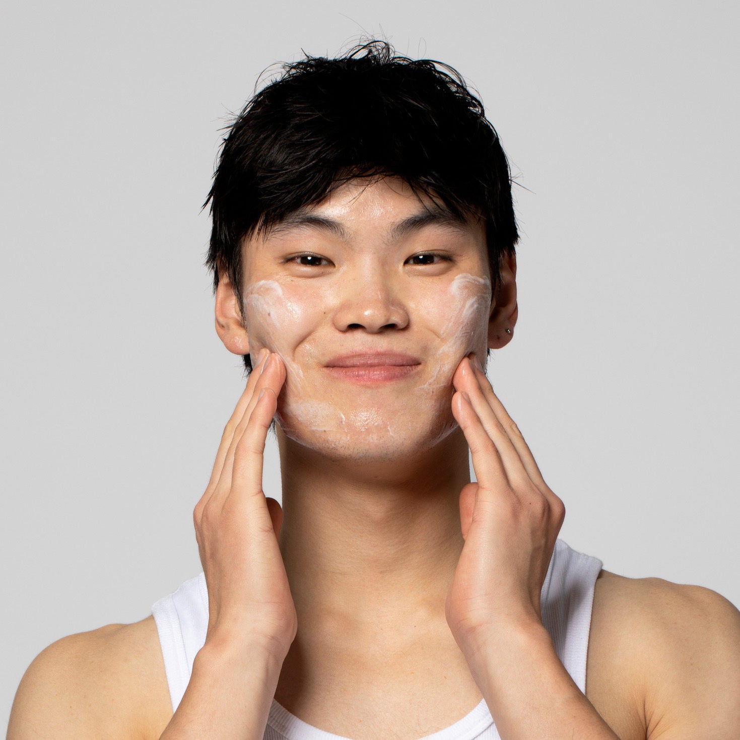 Junger Mann mit weißem Shirt wäscht sein Gesicht, lächelt leicht und hat Schaum auf den Wangen.