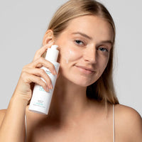 Frau trägt Hautpflegeprodukt aus weißer Flasche auf ihr Gesicht auf, mit einem Lächeln und leuchtendem Teint.